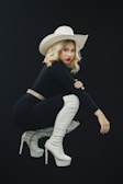 A woman with blonde hair is posing in a squatting position. She is wearing a large white cowboy hat, a black outfit, and white high-heeled thigh-high boots. Her makeup includes striking red lipstick, and she has a confident expression.