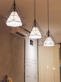 Three geometric pendant lamps with black wireframes hang from the ceiling, emitting a warm light. A wall-mounted air conditioner is visible on the left. Below the lamps, a wall has a quote from Aristotle about education. Neutral-colored blinds cover a portion of the background.