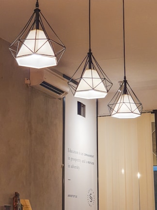Three geometric pendant lamps with black wireframes hang from the ceiling, emitting a warm light. A wall-mounted air conditioner is visible on the left. Below the lamps, a wall has a quote from Aristotle about education. Neutral-colored blinds cover a portion of the background.