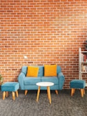 A living room setting features a blue sofa with two orange cushions and matching blue ottomans. In front of the sofa, there's a small round white table. The backdrop is a brick wall, and to the right, a bookshelf partially filled with books is visible. A potted plant is placed to the left of the sofa.