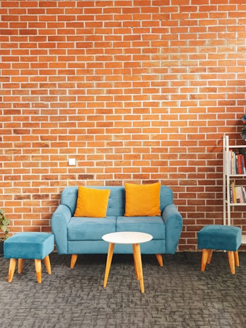A living room setting features a blue sofa with two orange cushions and matching blue ottomans. In front of the sofa, there's a small round white table. The backdrop is a brick wall, and to the right, a bookshelf partially filled with books is visible. A potted plant is placed to the left of the sofa.