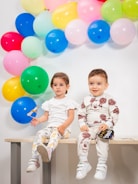 Two young children are sitting on a bench in front of a colorful arrangement of balloons. The child on the left is wearing a white shirt and pants with animal prints, holding a small sign. The child on the right is dressed in a matching outfit with a cartoon character print, also holding a small prop shaped like a speech bubble.