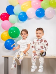 Two young children are sitting on a bench in front of a colorful arrangement of balloons. The child on the left is wearing a white shirt and pants with animal prints, holding a small sign. The child on the right is dressed in a matching outfit with a cartoon character print, also holding a small prop shaped like a speech bubble.