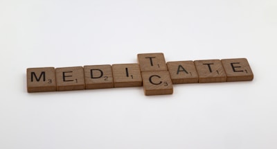 Wooden Scrabble tiles spell out the words 'MEDICATE' and 'MEDITATE', intersecting at the 'T'. Each tile displays a letter with an associated numerical value, typical of Scrabble games. The background is plain and minimalistic.
