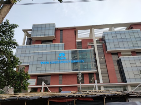 The image features the facade of a modern office building with a clean, geometric design. It is constructed with a combination of red brick and large glass panels, and there is a prominently displayed sign for Tata Consultancy Services in blue. Trees partially frame the view, and a lamppost is visible at the foreground.