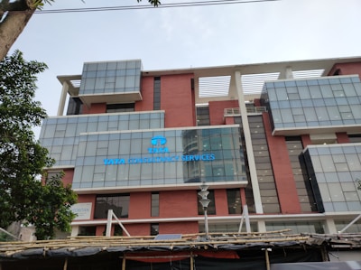 The image features the facade of a modern office building with a clean, geometric design. It is constructed with a combination of red brick and large glass panels, and there is a prominently displayed sign for Tata Consultancy Services in blue. Trees partially frame the view, and a lamppost is visible at the foreground.