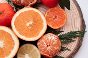Several citrus fruits, including oranges, tangerines, and a lemon, are placed on a wooden surface. Some fruits are sliced in half, showing their juicy interiors, while others are whole or partially peeled. Green leaves and small branches are also present, adding a natural touch to the arrangement.