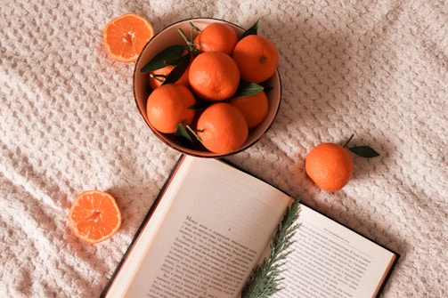 a bowl of oranges and an open book on a bed