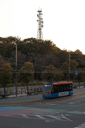 A tram traveling on a road with SOLATO branding visible on the side. The background is filled with dense trees and a tall communication tower with several antennas and dishes. The sky appears to be overcast or at dusk due to the lighting.