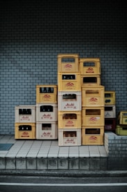 A stack of yellow and white crates against a blue brick wall, some filled with dark bottles. The crates are labeled with various brand names, adding a sense of industrial or commercial setting.