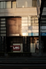 A building facade features a mix of windows and signage, displaying a blend of reflective and opaque surfaces. Large signs with Japanese text are visible, along with labels indicating EC and Mitsui Building. The architecture includes tiled patterns and metallic elements with sunlight casting shadows across the surface.