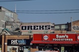 A cityscape with buildings featuring graffiti and signage. The building in the center has the word 'OREKS' prominently painted on it. Below, there are storefronts including 'Sam's Food Stores' and 'pet valu,' with signs for an ATM and other storefront advertisements. Power lines are visible crossing the scene.