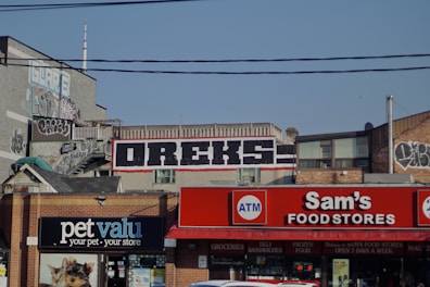 A cityscape with buildings featuring graffiti and signage. The building in the center has the word 'OREKS' prominently painted on it. Below, there are storefronts including 'Sam's Food Stores' and 'pet valu,' with signs for an ATM and other storefront advertisements. Power lines are visible crossing the scene.