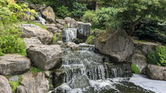 A close-up of a handcrafted ornamental waterfall with smooth stones and gentle water flow, surrounded by lush greenery.