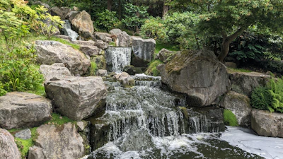 A close-up of a handcrafted ornamental waterfall with smooth stones and gentle water flow, surrounded by lush greenery.
