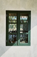 An old-fashioned window with a dark green frame and diamond-patterned glass, set in a plain, off-white wall. The glass reflects some outdoor elements like trees and a fence.