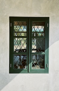 An old-fashioned window with a dark green frame and diamond-patterned glass, set in a plain, off-white wall. The glass reflects some outdoor elements like trees and a fence.