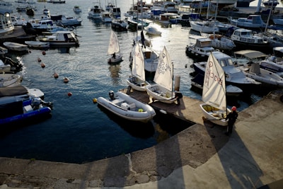 A marina filled with various boats, including small sailboats and motorboats, floating on calm water. The light from the setting sun casts long shadows and reflects on the water surface. Several individuals are preparing the small sailboats along the dock.