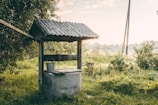 A rustic water well with a wooden roof, surrounded by lush greenery and a serene rural landscape. The setting sun casts a warm glow over the scene, enhancing the peaceful ambiance.