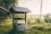 A rustic water well with a wooden roof, surrounded by lush greenery and a serene rural landscape. The setting sun casts a warm glow over the scene, enhancing the peaceful ambiance.