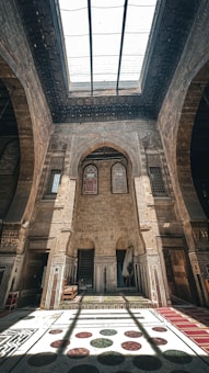 A grand interior of an ancient building with intricate stone carvings and large arches. Sunlight filters through a large glass skylight, casting shadows on a decorative marble floor. The walls are adorned with ornate details and there are traditional wooden furniture pieces.
