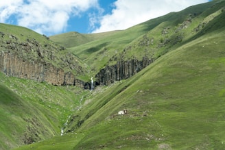 A serene mountain landscape with lush green hills and a small waterfall cascading down the rocky terrain. A solitary house is nestled among the hills, surrounded by the vast expanse of nature under a bright blue sky with scattered clouds.