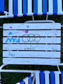 A white slatted chair with various hand-drawn messages and doodles, including blue and pink stars, and the Olympic rings drawn in black, blue, yellow, green, and red. The word 'Olympics' is written beneath the rings. The chair is positioned on a lawn with grass visible in the background.