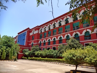 A large, multi-story red brick building with numerous arched windows and white accents sits surrounded by lush green trees and shrubs. The clear blue sky above and the paved courtyard in front enhance the bright and serene atmosphere.