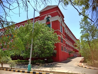 A large, red brick building with white accents features prominently, surrounded by greenery. The architecture includes numerous windows and a distinct arched design near the roof. The building is bordered by trees and a paved road with curbs painted in black and yellow stripes.