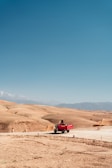 A red three-wheeled vehicle travels along a dirt road in a vast, arid landscape with rolling sandy hills and distant mountains under a clear blue sky.