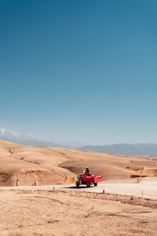 A red three-wheeled vehicle travels along a dirt road in a vast, arid landscape with rolling sandy hills and distant mountains under a clear blue sky.