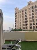 A tall beige building with multiple windows stands next to an outdoor area featuring a lounging chair on a patch of artificial grass. In the background, modern skyscrapers are visible under a partly cloudy sky.