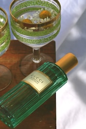 A green perfume bottle with a gold cap labeled 'Gucci Mémoire D'une Odeur' rests on a wooden surface. Beside it are two elegant glasses with gold and green decorative rims, containing floating white daisy flowers.
