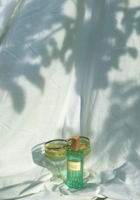 A perfume bottle with a greenish hue and gold cap is placed next to two green and gold detailed wine glasses. They sit on a white fabric that has shadows of leaves cast across it, creating an elegant and serene setting.
