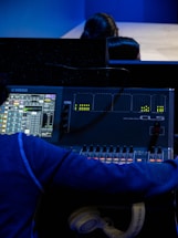 A person is operating a digital audio mixing console with a display showing various audio levels and controls. The console is branded Yamaha, and there are several adjustable faders and knobs. The individual is wearing a blue hoodie and has a pair of white headphones resting near the console.