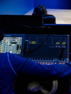 A person is operating a digital audio mixing console with a display showing various audio levels and controls. The console is branded Yamaha, and there are several adjustable faders and knobs. The individual is wearing a blue hoodie and has a pair of white headphones resting near the console.