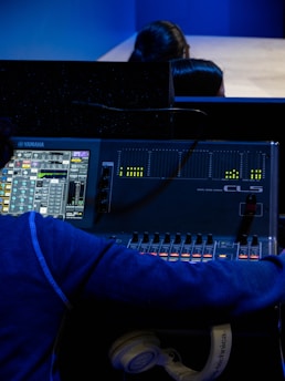 A person is operating a digital audio mixing console with a display showing various audio levels and controls. The console is branded Yamaha, and there are several adjustable faders and knobs. The individual is wearing a blue hoodie and has a pair of white headphones resting near the console.