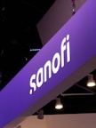A prominently displayed sign with the name 'sanofi' is illuminated against a purple background. The sign seems to be part of an exhibition or conference setup, with multiple spotlights hanging from the ceiling.