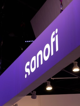 A prominently displayed sign with the name 'sanofi' is illuminated against a purple background. The sign seems to be part of an exhibition or conference setup, with multiple spotlights hanging from the ceiling.