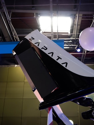 A close-up view of a sleek, modern machine or device with the word 'Zapata' prominently displayed on it. The setting appears to be indoors with industrial-style lighting and metal framework visible on the ceiling. The device features a combination of black and white colors, possibly part of a larger structure or vehicle.