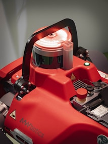 A close-up view of a red robotic device featuring a prominent handle on top, several controls, and buttons. The design includes caution labels and the branding 'AMYbotics'. The lighting accentuates the machine's surfaces, emphasizing its industrial nature.