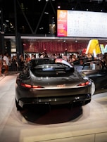 A sleek, futuristic Audi car is displayed in a well-lit exhibition or showroom. The vehicle's streamlined design features prominent taillights and a glossy finish. In the background, a diverse crowd of people are gathered, some taking photos of the car. Above them is a large digital screen displaying logos and banners related to technology and partnerships, suggesting a tech event or auto show.