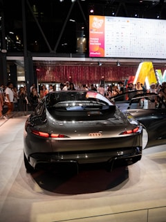 A sleek, futuristic Audi car is displayed in a well-lit exhibition or showroom. The vehicle's streamlined design features prominent taillights and a glossy finish. In the background, a diverse crowd of people are gathered, some taking photos of the car. Above them is a large digital screen displaying logos and banners related to technology and partnerships, suggesting a tech event or auto show.