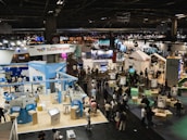 A large, bustling exhibition hall with numerous booths and displays from various companies, some of which have recognizable brand logos. There is a significant number of people walking around, engaging with displays, and interacting with exhibitors. The space is well-lit with overhead lighting, and there are multiple banners and signs throughout the area.