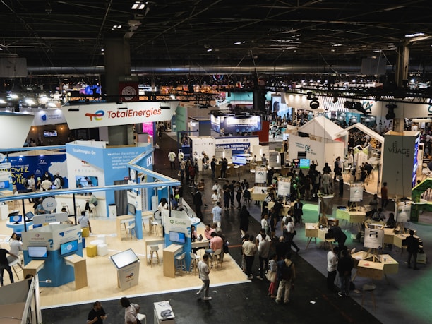 A large, bustling exhibition hall with numerous booths and displays from various companies, some of which have recognizable brand logos. There is a significant number of people walking around, engaging with displays, and interacting with exhibitors. The space is well-lit with overhead lighting, and there are multiple banners and signs throughout the area.