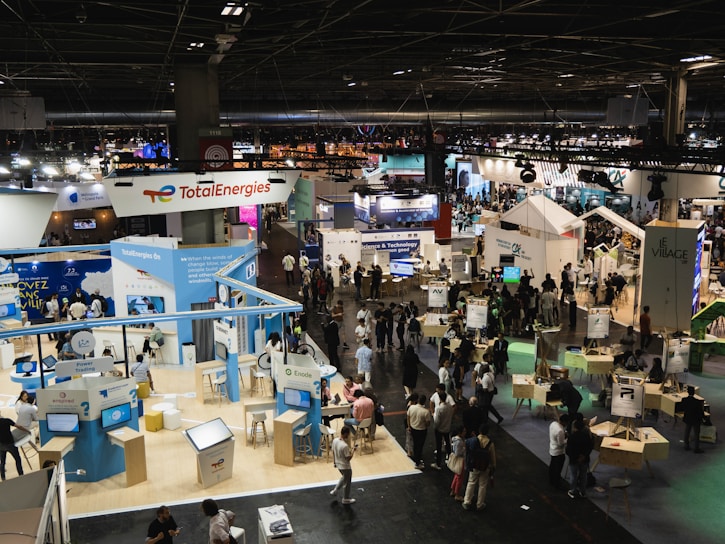 A large, bustling exhibition hall with numerous booths and displays from various companies, some of which have recognizable brand logos. There is a significant number of people walking around, engaging with displays, and interacting with exhibitors. The space is well-lit with overhead lighting, and there are multiple banners and signs throughout the area.