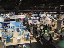 A large, bustling exhibition hall with numerous booths and displays from various companies, some of which have recognizable brand logos. There is a significant number of people walking around, engaging with displays, and interacting with exhibitors. The space is well-lit with overhead lighting, and there are multiple banners and signs throughout the area.