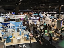 A large, bustling exhibition hall with numerous booths and displays from various companies, some of which have recognizable brand logos. There is a significant number of people walking around, engaging with displays, and interacting with exhibitors. The space is well-lit with overhead lighting, and there are multiple banners and signs throughout the area.