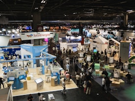A large, bustling exhibition hall with numerous booths and displays from various companies, some of which have recognizable brand logos. There is a significant number of people walking around, engaging with displays, and interacting with exhibitors. The space is well-lit with overhead lighting, and there are multiple banners and signs throughout the area.