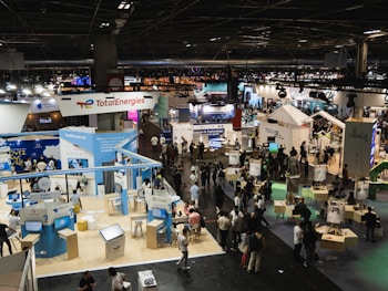 A large, bustling exhibition hall with numerous booths and displays from various companies, some of which have recognizable brand logos. There is a significant number of people walking around, engaging with displays, and interacting with exhibitors. The space is well-lit with overhead lighting, and there are multiple banners and signs throughout the area.